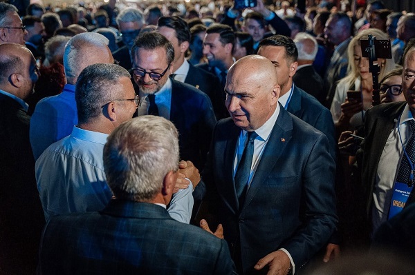Ilie Bolojan vesel, imbracat cu costum bleumarin si camasa alba, strangandu-i mana unui barbat cu camasa alba si cu ochelari, in timp ce este inconjurat de sustinatori si de jurnalisti la Congresul Extraordinar PNL