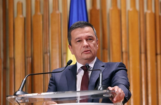 Sorin Grindeanu imbracata cu sacou albastru, camasa alba si cravata visiunie stand in fata unui pupitru cu microfoane, intr-o sala cu peretii captusiti cu lemn, avand in spatele sau un steag albastru