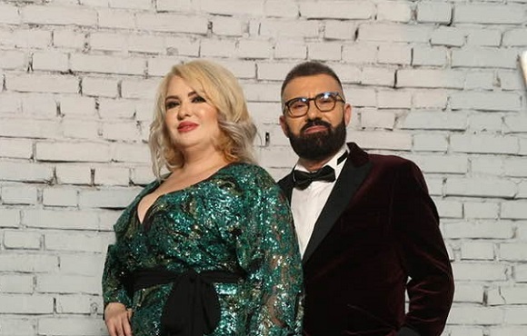 Viorica de la Clejani zambitoare, imbracata cu rochie verde din material stralucitor, cu cordon negru, alaturi de sotul ei, Ionita de la Clejani, purtand ochelari, imbracat cu sacou visiuniu de catifea, cu camasa alba si cu papion negru, amandoi stand rezemati de un perete de caramida alba