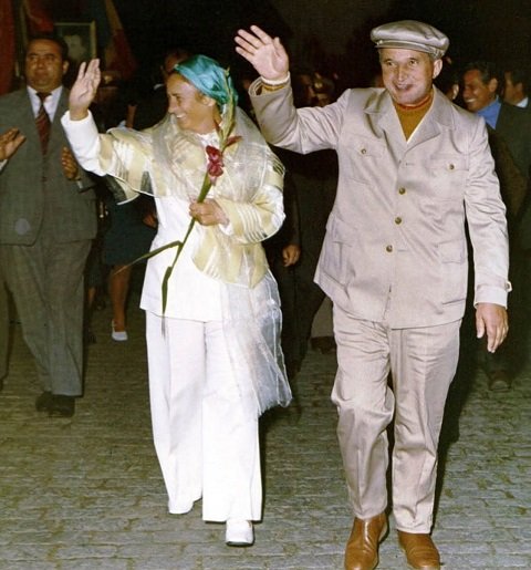 Elena Ceausescu imbracata intr-un costum alb cu pantaloni, cu o basma bleu-turcoaz pe cap, cu o esarfa transparenta pe umeri, tinand in mana o floare rosie si cu cealalta facand cu mana, in timp ce in spatele ei se afla un barbat in costum gri, iar langa ea se afla sotul ei, Nicolae Ceausescu, imbracat cu costum gri deschis, cu pulover maro pe gat si purtand sapca asortata, in timp ce face si el cu mana oamenilor din multimea de pe strada