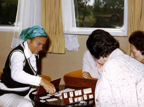 Elena Ceausescu imbracata in alb, cu vesta negru cu alb, cu basma albastra pe cap, jucand rummy cu cateva femei intr-un comportiment de tren