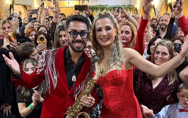 Claudia Puican vesela, imbracata cu rochie rosie decoltata, facand cu mana, in timp ce alaturi de ea se afla sotul ei, saxofonistul Armin Nicoara, imbracat cu sacou rosu cu strasuri pe un umar si cu camasa alba, si el ridicand bratul in timp ce impreuna s-au aflat la o petrecere de Revelion alaturi de numeroase persoane 