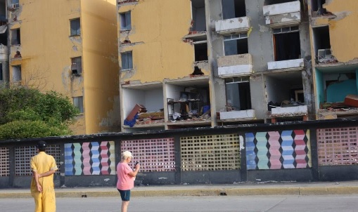blocuri vopsite in galben si gri grav avariate dupa cutremurele din Venezuela, in fata blocurilor fiind garduri colorate si persoane care fac fotografii si se uita la dezastrul provocat de cutremure