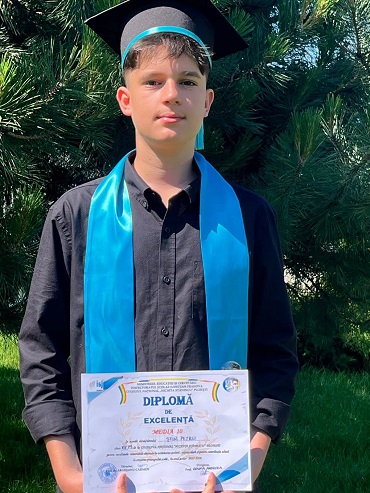 Petru Știr, elevul din Prahova care a avut media 10 la Evaluarea Nationala, purtand toca neagra pe cap si roba de absolvent neagra cu panglica turcoaz, tinand in maini o diploma, stand cu spatele la un molid