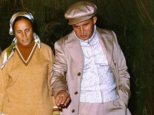Elena Ceausescu imbracata cu pulover maroniu, cu basma cu nuante de maro pe cap, in timp ce priveste in jos, stand langa sotul ei, Nicolae Ceausescu, imbracat cu pulover pe gat si sacou gri-bej, cu sapca pe cap, intr-un cadru nocturn, in perioada in care au vizitat Delta Dunarii