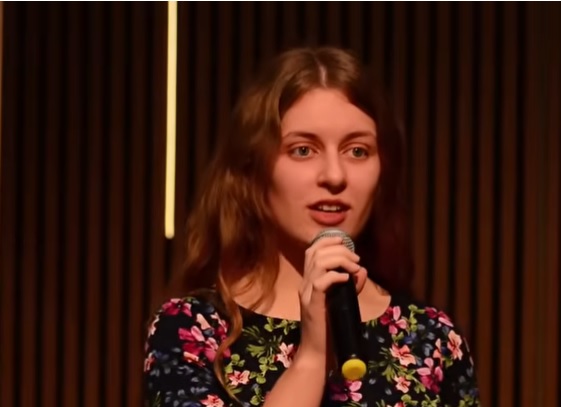 Eliana, fiica cea mare a soților Bodnariu, imbracata cu rochie neagra cu flori mari, cu maneca scurta, tinand in mana un microfon in timp ce vorbeste pe o scena