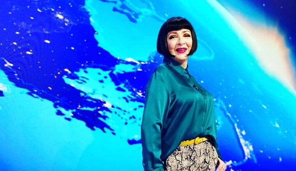 Neti Sandu zambitoare, imbracata cu bluza turcoaz din material lucios, cu pantaloni cu imprimeu piele de sarpe si curea galbena, in spatele ei fiind un decor din studiourile de la PRO TV