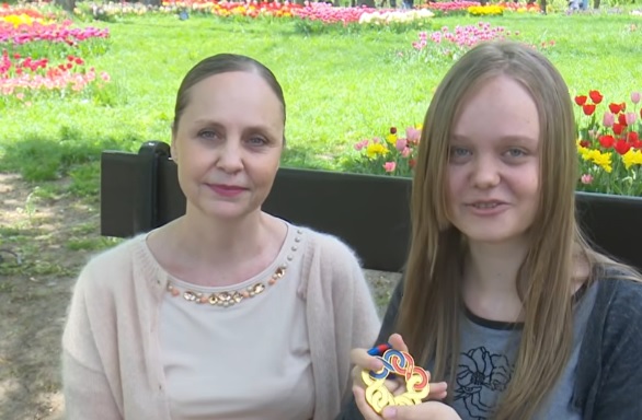 captură video YouTube TVR Craiova, Carina Viespescu imbracata cu tricou gri, tinand in mana o medalie de aur, in timp ce sta pe o banca alaturi de mama ei, imbracata cu pulover si bluza bej, amandoua aflandu-se intr-un parc primavara