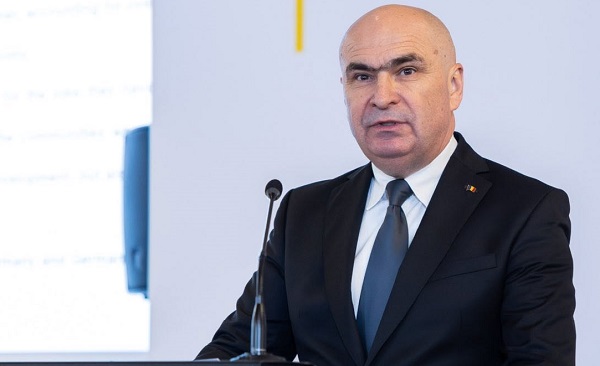 premierul interimar Ilie Bolojan imbracat cu camasa alba, sacou bleumarin si avand cravata albastra, in timp ce sta in fata unui pupitru cu microfoane, tinand un discurs, intr-un spatiu interior cu pereti albi si cu proiector