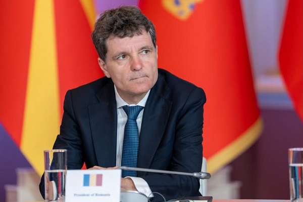 președintele Nicusor Dan ganditor si abatut, imbracat cu camasa alba, cu sacou bleumarin si avand cravata albastra, in timp ce sta la o masa cu microfoane la o reuniune, avand in spatele sau diverse steaguri cu rosu si in fata lui un pahar cu apa si un biletel cu drapelul Romaniei