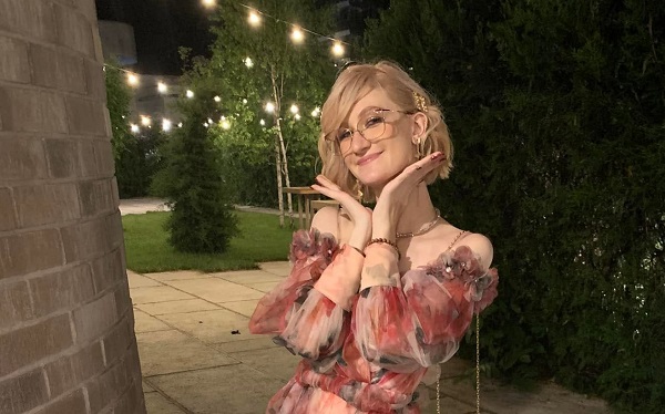 Anastasia Iris Piron vesela, purtand ochelari, imbracata cu o rochie de voal inflorata in nuante de roz, tinandu-si mainile sub barbie ca sa pozeze intr-un cadru nocturn, cu o gradina luminata si cu o cladire de piatra