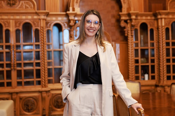 Diana Buzoianu vesela, purtand ochelari, imbracata cu bluza neagra si costum cu pantaloni bej cu dungi fine maro, stand sprijinita de un scaun de lemn intr-o incapere cu mobilier de lemn