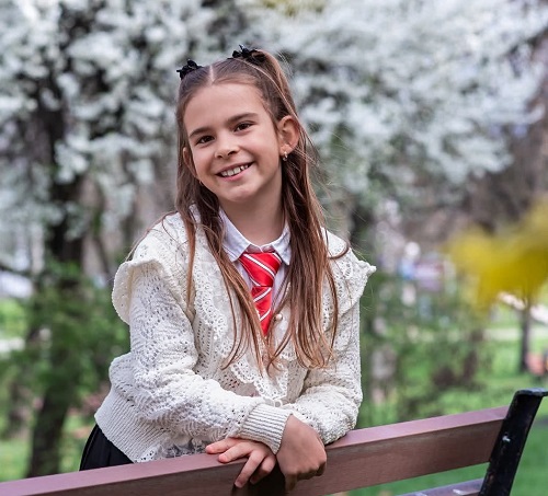 Raisa, fetita de 9 ani care a murit in urma accidentului de la Cluj, vesela, imbracata cu puloveras alb, camasa alba si cravata rosie cu dungi albe, stand cu bratele sprijinite de o banca, in timp ce se afla in aer liber, intr-un cadru cu copaci infloriti primavara