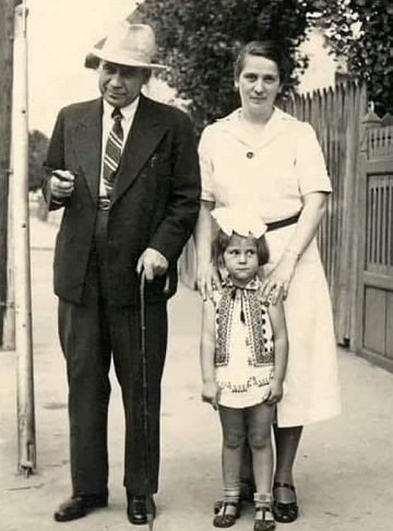 fotografie alb-negru, medicul Dumitru Bagdasar imbracat cu costum, purtand palarie pe cap, tinand intr-o mana o tigara si in alta un baston, alaturi de el aflandu-se sotia lui, Florica Bagdasar, imbracata cu rochie, care tine mainile pe umerii fiicei lor, Alexandra, imbracata cu vesta inspirata din ilicul popular, cu funda pe cap, toti trei aflandu-se pe o strada, langa un gard