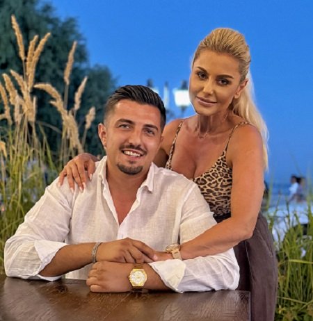 Emilia Ghinescu zambitoare, imbracata cu top animal print, in timp ce isi tine mainile pe umerii partenerului sau, Viorel Neagoe, zambitor, imbracat cu camasa alba, in timp ce Viorel sta pe scaun la o masa intr-un spatiu cu iarba, in apropiere de malul marii