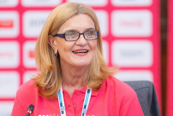 Elisabeta Lipă veselă, purtand ochelari cu rame negre, imbracata cu tricou roz intens, in timp ce sta pe scaun in fata unui microfon la o conferinta