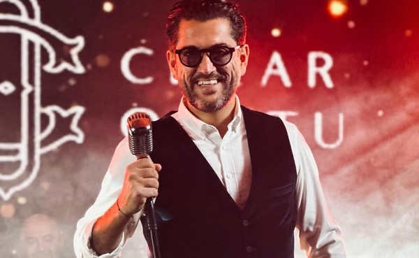 Cezar Ouatu vesel, purtand ochelari negri, imbracat cu camasa alba si vesta neagra de catifea, in timp ce tine cu mana un microfon la un concert al sau