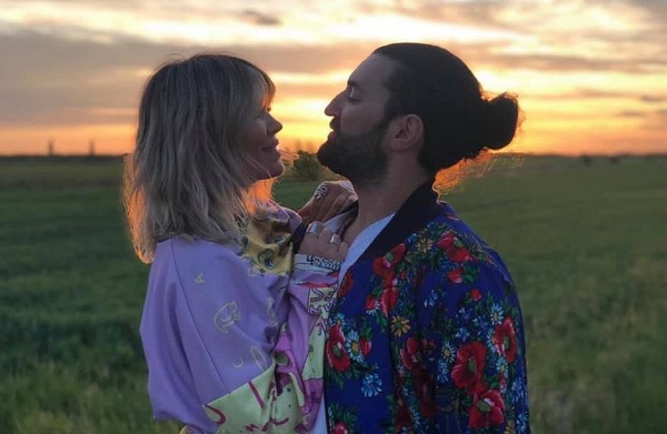 Gina Pistol imbracata cu o bluza cu lila si cu galben pai, cu maneca lunga, in timp ce se uita zambind in ochii partenerului ei, Smiley, imbracat cu o jacheta albastra inflorata, amandoi aflandu-se pe un camp la apusul soarelui