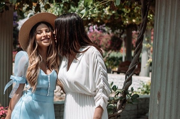 Valerie Lungu fericita, imbracata cu rochie lunga bleu, avand pe cap o palarie, langa ea stand mama ei, Victoria Lungu, imbracata intr-o rochie alba, amandoua aflandu-se intr-o gradina