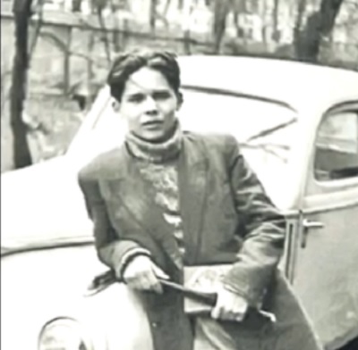 fotografie alb-negru, poetul Nicolae Labis imbracat cu pulover pe gat si sacou, stand rezemat de o masina langa un spatiu cu copaci