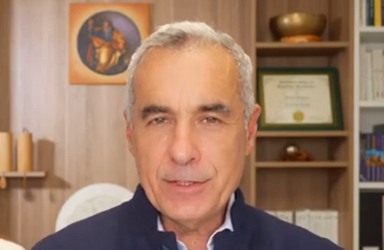 captură video YouTube, Călin Georgescu imbrăcat cu cămașă alba si pulover albastru, in timp ce se filmeaza pentru propriul canal de YouTube in timp ce se afla in locuinta sa, in spatele sau putandu-se observa diverse piese de mobilier