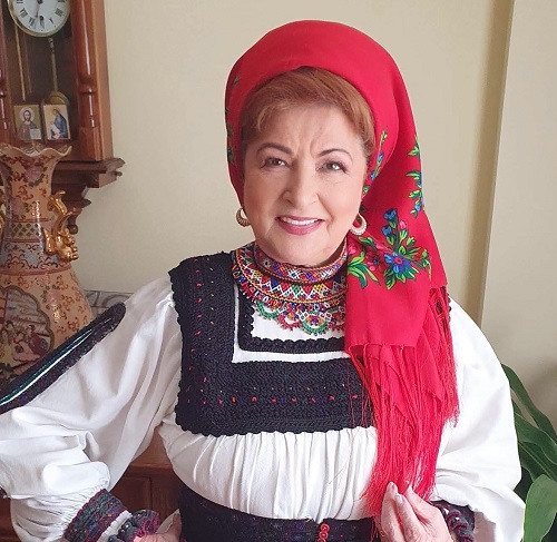 Maria Petca Poptean zambitoare, cu basma rosie inflorata, imbracata cu port traditional, in spatele ei aflandu-se un ceas cu pendula,o vaza si alte obiecte dintr-un spatiu interior