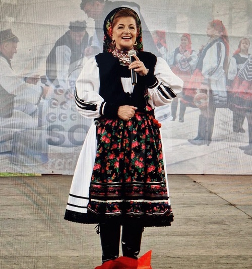 Maria Petca Poptean cu basma rosie, imbracata cu port din Tara Oasului, purtand cizme negre, tinand microfonul in mana in timp ce se afla pe o scena