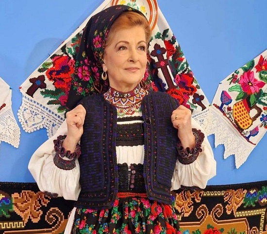 Maria Petca Poptean ganditoare, cu batic negru inflorat, imbracata in port popular, tinandu-si mainile pe ilic, in spatele ei aflandu-se un perete albastru si stergare brodate
