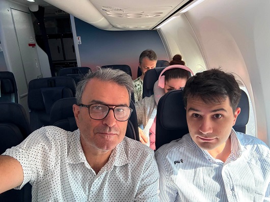 Lucian Mindruta purtand ochelari, imbracat cu camasa cu maneca scurta alba cu imprimeu marunt, alaturi de fiul sau cel mare, Mihai, imbracat cu camasa alba cu dungi,amandoi stand pe scaun intr-un avion, in spatele lor observandu-se pasageri