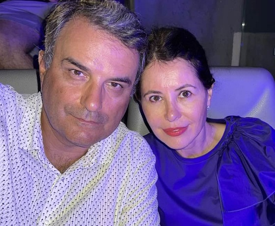 Lucian Mindruta imbracat cu camasa alba cu imprimeu marunt, stand pe o canapea de piele gri alaturi de sotia lui, Ioana Mindruta, imbracata intr-o tinuta albastra din material satinat