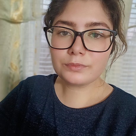 Ioana, tanara ucisa de sot in Vrancea, imbracata in bleumarin inchis, avand parul strans si ochelari cu rame negre, stand cu spatele la o fereastra cu storuri albe si perdea