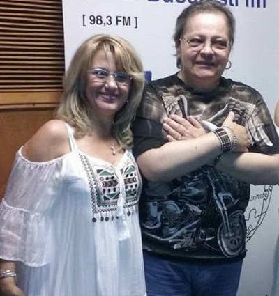 Paula Iorga vesela, purtand ochelari, imbracata cu rochie alba cu broderie, cu umerii dezgoliti, alaturi de sotul ei, Leo iorga, purtand ochelari, imbracat cu tricou negru cu imprimeu si blugi, tinand bratele incrucisate pe piept, amandoi aflandu-se intr-un studio de radio