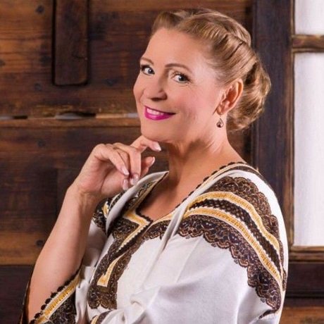 Matilda Pascal Cojocarita cu parul strans, zambitoare, imbracata in costum popular si tinandu-si mana sub barbie, in timp ce sta intr-un cadru rustic cu mult lemn