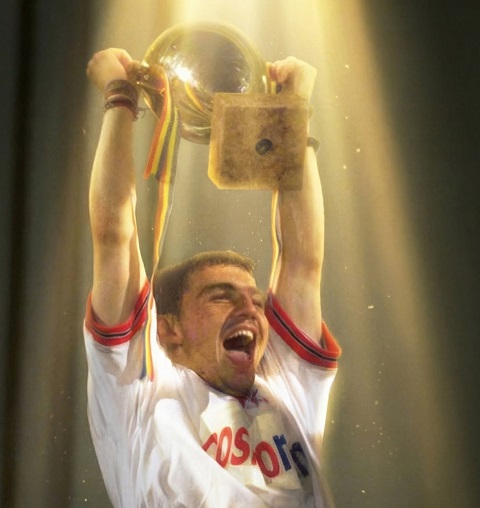 Cătălin Hîldan fericit, imbrăcat in echipament sportiv alb cu rosu, ridicand deasupra capului o cupă, fiind invaluit intr-o lumina aurie