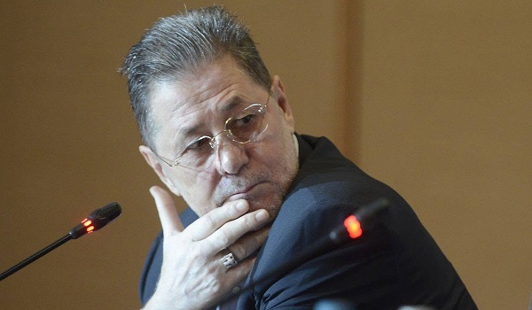Cornel Dinu ganditor, purtand ochelari, imbracat cu costum negru, in timp ce isi duce mana la barbie cand sta pe scaun, la o masa, in fata lui fiind un microfon