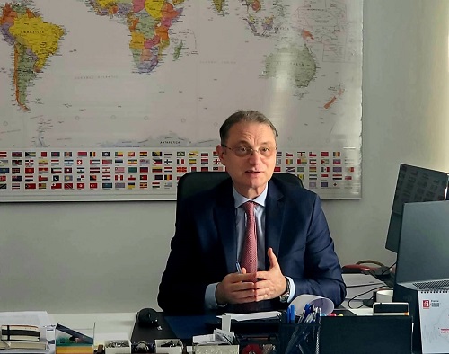 Luca Niculescu purtand ochelari, imbracat cu costum bleumarin, avand camasa bleu si cravata visinie, in timp ce sta biroul sau, avand in spate o harta a lumii pusa pe perete 