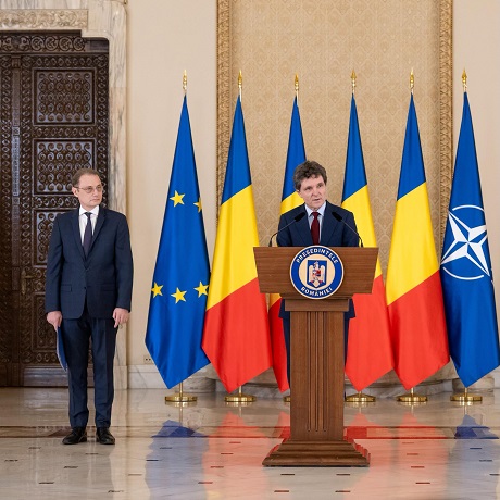 Nicusor Dan imbracat cu costum bleumarin, stand la pupitru cu microfon la Administratia Prezidentiala in timp ce anunta nominalizarea lui Luca Niculescu pentru functia de premier si Luca Niculescu, premierul desemnat, purtand ochelari, imbracat cu costum bleumarin, stand cu spatele o usa de lemn masiv si la steagurile aflate in sala de la Administratia Prezidentiala
