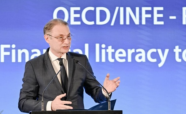 Luca Niculescu purtand ochelari, imbracat cu costum negru, avand camasa crem si cravata neagra, stand in fata unui pupitru cu microfon si tinand un discurs intr-o conferinta a OCDE