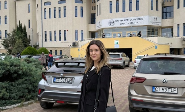 Melisa Seic, tanara din judetul Constanta care a murit la 19 ani, zambitoare, imbracata cu bluza alba si sacou negru, avand geanta pe umar, in timp ce sta in preajma a doua masini argintii in parcarea de la Facultatea de Medicina a Universitatii Ovidius din Constanta
