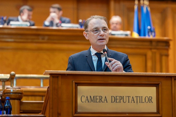 Luca Niculescu purtand ochelari, imbracat cu costum bleumarin, camasa alba si purtand cravata bleumarin, incercand sa-si regleze microfonul in timp ce statea la pupitru in Parlament, la Camera Deputatilor, pentru a-si susține discursul, in timp ce în spatele sau se afla mai mulți membri ai Parlamentului