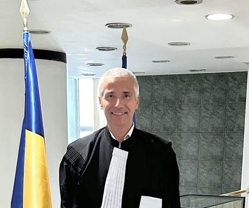 Radu Marinescu, Ministrul Justitiei, zambitor, imbracat in roba neagra, stand langa doua steaguri intr-o incapere