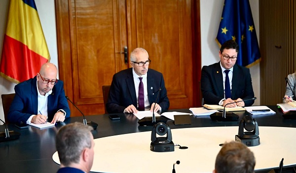 Radu Marinescu cu ochelari, imbracat cu camasa alba, sacou albastru si cravata violet, stand pe scaun si coordonand prima sedinta de la Ministerul Justitiei, intr-o sala cu steagul Romaniei si cu drapelul Uniunii Europene, in prezenta mai multor persoane