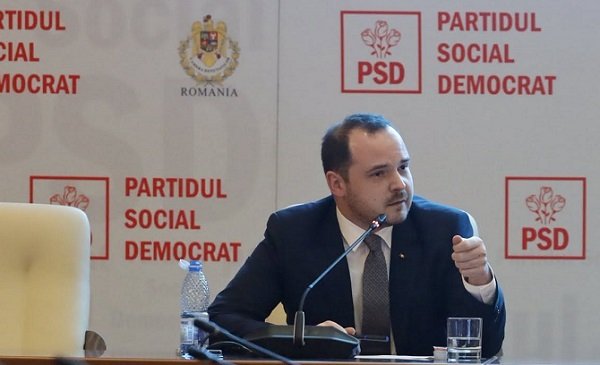 Alexandru Rogobete, ministrul Sanatatii, imbracat cu sacou bleumarin si camasa albastra, stand la o masa in fata unui microfon, in timpul unei sedinte din cadrul Partidului Social Democrat