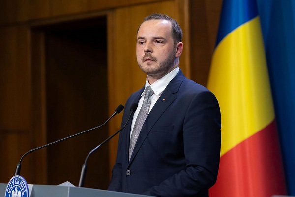 Alexandru Rogobete, ministrul Sanatatii, imbracat cu costum bleumarin, camasa alba si purtand cravata albastra, stand la pupitrul cu sigla Guvernului Romaniei, la microfon, avand in spatele sau steagul Romaniei si o usa de lemn, tinand un discurs 