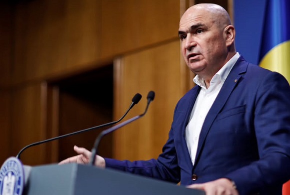 Ilie Bolojan imbracat cu sacou bleumarin si camasa albastra, stand la pupitru, in fata microfoanelor si dand o declaratie la Guvern, in spatele sau fiind o usa de lemn si drapelul Romaniei