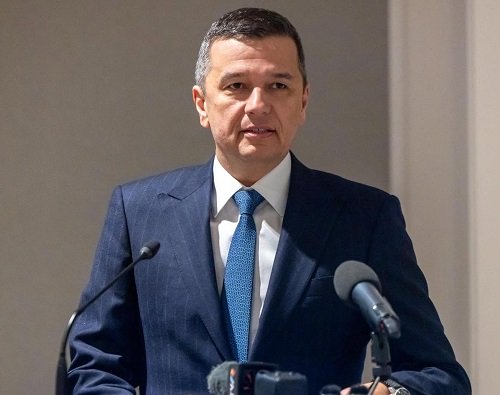 Sorin Grindeanu imbracat cu sacou bleumarin, camasa alba si purtand cravata albastra, stand la un pupitru si vorbind la microfon intr-un spatiu in aer liber, intr-o zi insorita
