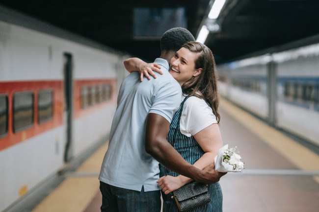 Cuplu care se imbratiseaza pe peron de metrou, ea tine un buchet de flori in mana, primit de la el