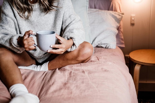 Femeie care sta pe pat, imbracata in pulover si sosete si bea ceai din ceasca