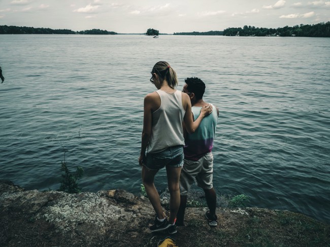 Cuplu fotografiat de la spate care priveste in jos, intr-un lac, de pe o stanca