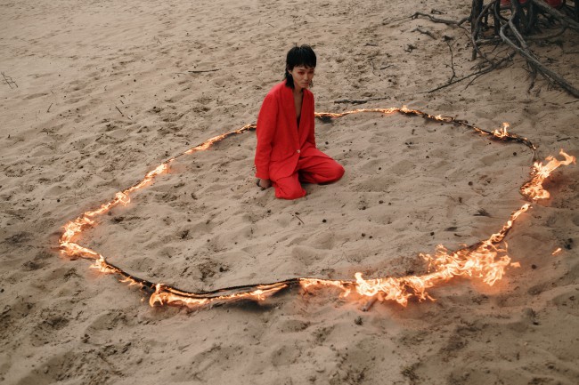 Femeie bruneta in haine largi rosii care sta pe nisip intr-un cerc de foc
